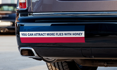 Horizontal striped red, white and blue, bumper sticker on lower left rear bumper of car. It reads You Can Attract More Flies With Honey.
